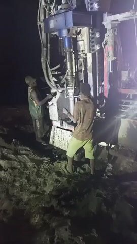 Two men are operating a large drilling rig at night. The rig is off-white with dark blue accents and is surrounded by muddy ground. One man is wearing a tank top and dark shorts, while the other wears a patterned shirt and light green shorts, both also wear boots. They are both focused on the machinery, seemingly adjusting controls and overseeing the drilling process. The surrounding environment is dark, lit only by the lights of the machinery.
