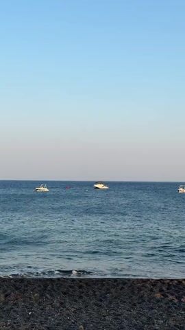 A vertical video shows a tranquil ocean scene with a dark pebble beach in the foreground. The calm blue water extends to the horizon, where it meets a clear, pale blue sky. Three white boats are visible on the water. One small boat is on the left, closer to the shore. Another slightly larger boat is in the middle, with a red buoy nearby. A third boat is on the right, further away. The waves are gentle, lapping at the edge of the beach, creating a peaceful and serene atmosphere.