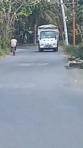 A white truck is driving down a paved road. To the left of the truck, a person on a bicycle is also traveling down the road. Trees and foliage line the sides of the road. The sky is overcast.