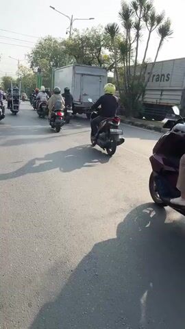 The video shows a line of motorcycles on a paved road, with a large truck in the background. The sky is clear and bright, suggesting a sunny day. The motorcycles are all moving in the same direction, and the riders are wearing helmets. Some of the riders have passengers. The road appears to be in a city or suburban area, with trees and buildings visible in the background. The overall atmosphere is one of everyday urban life, with the sound of traffic and motorcycles filling the air.