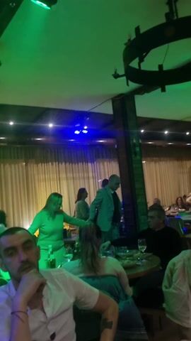 The video shows a group of people dining and dancing in a restaurant setting.  The restaurant is lit with colored lights that change from green to pink to blue.  There are multiple tables with people seated at them, eating and drinking. Several people are standing and dancing, including a man in a light blue blazer. A circular chandelier hangs from the ceiling. The room is decorated with light-colored curtains and wooden accents. The camera is held by someone at a table.  Some of the people at the table are looking at the dancers and others are looking at the camera.