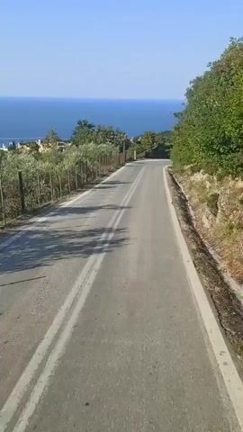 The video shows a paved road with double white lines, curving to the right. The road is bordered by a wire fence and vegetation on the left, and a rocky slope with trees and bushes on the right. In the background, the blue sea stretches to the horizon under a clear blue sky. There are scattered trees and some buildings visible on the hillside overlooking the sea. The lighting suggests it is daytime, likely morning or afternoon, with bright sunlight casting shadows. The overall scene evokes a sense of travel and scenic beauty, possibly in a coastal region of Greece. No people are visible, and no text is present in the video.