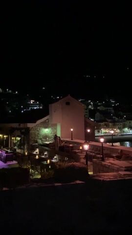 The video depicts an outdoor restaurant scene at night. The restaurant is illuminated by lamplight, candles, and purple ambient lighting. Tables and chairs are arranged on different levels. A white building with a light on it stands prominently in the middle of the frame. In the background, faint lights from other buildings on a hillside are visible. Some people are standing and talking on the upper level, near the white building. The camera slowly pans slightly to the left.