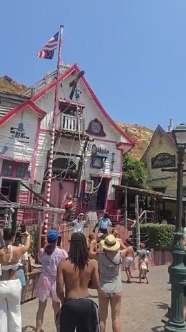 A group of people, many in swimwear, are gathered outdoors in what appears to be a themed village. In the center is a two-story, brightly painted white and pink building with a tilted appearance, resembling a pirate-themed structure. A flagpole with an American flag flies from the top of the building. Several individuals, seemingly performers dressed as pirates, are interacting with the crowd on a wooden deck in front of the building. Some audience members are holding up their phones to record. The environment suggests a sunny day, with a clear blue sky and hills in the background. Other themed buildings, including one with a sign that says "GENERAL STORE," are visible to the right.