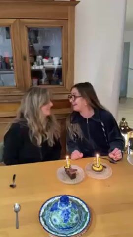 Two women, likely sisters, are celebrating a birthday at a wooden table. Each has a small dessert, likely a cupcake, with a lit candle. The woman on the right is wearing glasses and a dark blue zippered jacket. The woman on the left is also wearing a dark zippered jacket and has longer, lighter hair. They are both smiling and making gestures with their hands, seemingly dancing. A large wooden cabinet with glass doors stands behind them, filled with various objects. A spoon is on the table in front of each woman, and a decorative blue and white dish sits in the center. The background is a neutral-colored wall with what appears to be a staircase in the distance.