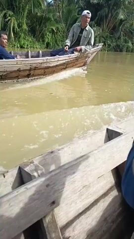 The video shows two men in a wooden boat on a muddy river. The man in the front is wearing a blue shirt and smiling. The man in the back is wearing a light-colored jacket and a white cap. He is looking towards the man in front and appears to be speaking. The boat is moving, creating a wake. The riverbanks are lined with lush green vegetation, including palm trees. The scene appears to be in a tropical environment. The camera is positioned in another boat traveling alongside the first boat, looking towards the two men.