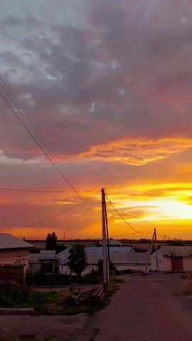 The video begins with a low-angle shot of a rural village at sunset. The sky is ablaze with vibrant hues of orange, yellow, and pink, with scattered clouds creating a dramatic effect. The foreground features a dirt road leading into the village, flanked by power lines and utility poles. Small houses and buildings are visible in the midground, some with red roofs. Trees dot the landscape, adding touches of green against the warm tones of the sky. As the video progresses, the camera pans slightly, revealing more of the village and the surrounding area. The lighting shifts subtly, intensifying the colors of the sunset. The final frames show a car driving along the road, further emphasizing the peaceful, everyday life in this rural setting. The overall impression is one of natural beauty and tranquility, capturing a serene moment as day transitions into night.