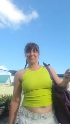 A woman with dark hair pulled into braids, wearing a lime green tank top and a white skirt, stands outdoors with a purple bag slung over her shoulder. She smiles and gestures with her hands. In the background, a blue sky with scattered clouds is visible, along with a white building and some greenery. Another person, partially visible, walks in the background.