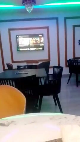 The video showcases the interior of a restaurant with modern, colorful lighting.  The camera pans upward, focusing on the ceiling which features multiple circular light fixtures with pink, blue, and green LED lighting. A chandelier with crystal accents hangs from the center of one of the light fixtures. The camera then pans down, showing dark tables and chairs in the foreground, and a TV screen on the wall in the background. The restaurant appears empty.