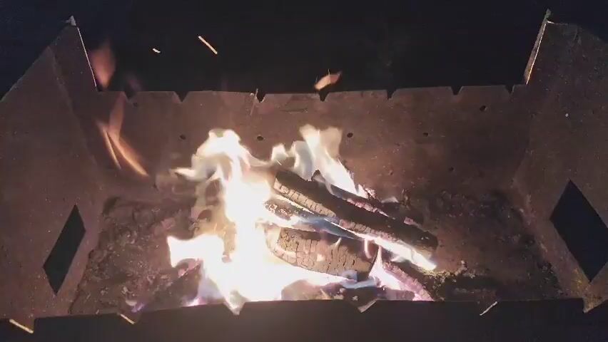 A fire burns in a metal brazier. The fire is mostly yellow and orange flames, with some blue at the base. There are several pieces of wood in the fire, some of which appear to be partially burnt. The brazier is dark metal and has a sawtooth edge around the top. The background is dark, suggesting the video was taken at night or in a dark environment. There are no people or other objects visible. The video is short, approximately 5 seconds long.