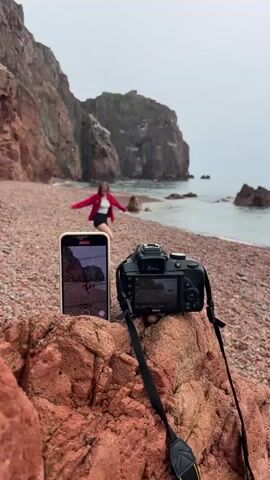 The scene is a rocky beach with red sand, set against a backdrop of large, rocky cliffs meeting the sea. The sky is overcast. In the foreground, a DSLR camera and a smartphone stand on a large reddish rock. Both devices are displaying the image of a young woman standing on the beach. The woman is wearing a red jacket and black shorts. She initially stands with her arms outstretched and then lowers them to her sides, posing. The camera and the phone are positioned to capture the scene, suggesting someone is taking pictures or videos of the woman.  The environment is rugged and natural, with the sound of waves audible.
