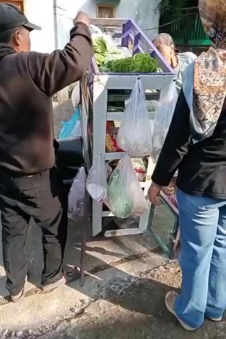 A vendor with a cart of produce is interacting with two customers. The vendor, a man wearing a black hooded sweatshirt and a black baseball cap, is on the left side of the cart, facing away from the camera. He is holding a bunch of leafy greens and placing them into a white plastic bag. A woman, wearing a light blue shirt and a patterned headscarf, is standing on the right side of the cart, facing the vendor. She is holding her left hand up and appears to be gesturing. Behind her, another woman is partially visible, wearing a black top and blue jeans, and a patterned headscarf. The cart is made of wood with purple accents and has shelves filled with vegetables and bagged items. The background shows a building with windows and a wall with some greenery. The ground is paved with dirt and some debris. The lighting suggests it is daytime.