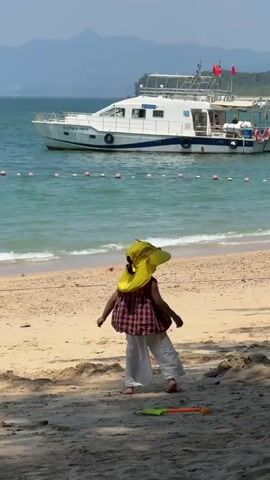 A young child wearing a yellow sun hat, a checkered shirt, and white pants stands on a sandy beach. The child turns their back to the camera and walks away. In the background, a white boat with blue trim is docked in the turquoise water. Waves gently lap the shore. A few small buoys are visible in the water, marking a boundary. The sand is light brown, and some dry leaves and shadows are on the ground in the foreground. A green and orange toy shovel lies on the sand near the child. The sky is clear and blue, and distant, hazy mountains are visible.