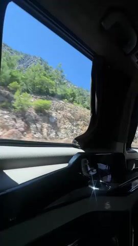 The video shows a view from inside a vehicle, looking out the passenger side window. The window is partially obscured by a dark curtain on the right side. Outside the window, there is a rocky mountainside with sparse green vegetation and trees. The sky is clear and blue. The interior of the vehicle is dark and features a shiny, black surface in the foreground with what appears to be a control panel. The vehicle appears to be in motion, as indicated by the slight blur of the scenery outside. There are no people visible in the video.