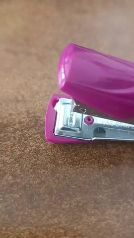 A close-up, static shot shows a pink stapler resting on a brown, textured surface. The stapler is open, revealing its metallic interior mechanism and a few staples. The lighting appears to be natural and diffused.