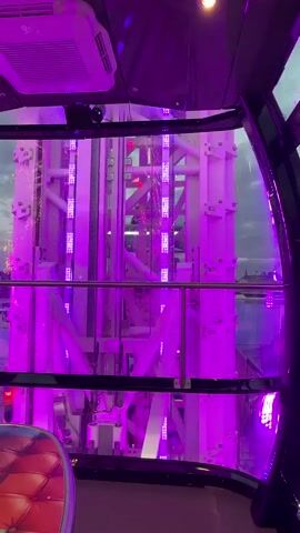 The video shows the interior of a gondola on a Ferris wheel, looking out at an amusement park at dusk. The gondola has purple LED lighting, and a padded seat with a colorful, quilted pattern is visible in the foreground. Outside, the park is illuminated with purple lights, and several rides, including a roller coaster, are visible. The sky is cloudy and a dim twilight. Raindrops are visible on the glass of the gondola.