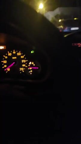 The video appears to be shot from the interior of a car at night, looking forward. The dashboard is visible with illuminated gauges, showing the speedometer needle in the 170-180 km/h range. A street light illuminates the scene outside, casting a glow on trees and houses. Other cars are visible in the distance, with their taillights illuminated. The video then briefly shows a close-up of a yellow line on the road, followed by a blurry shot of the rear of a car.