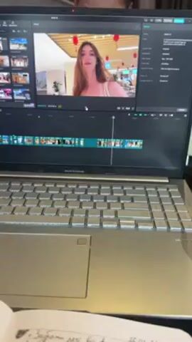 A laptop computer is open on a table, showing a woman in a white top on the screen within a video editing application. The video editing software has a timeline with many short video clips strung together. The woman on the screen is centered and looks toward the camera. There is what appears to be a book or stack of papers in the foreground, partially obscuring the bottom edge of the laptop. The laptop is an ASUS Vivobook.