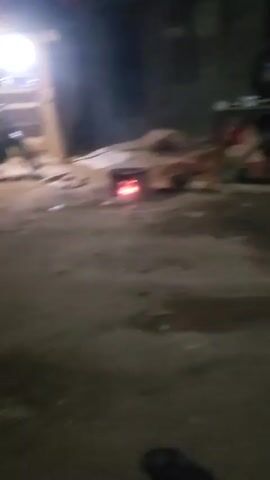 The video shows an outdoor scene at night, partially illuminated, with a white vehicle parked. As the camera moves, it focuses on a person sitting on a patterned mat. The person is wearing a red and white striped shirt with black pants and gestures toward the camera. Several other people appear in the background but are mostly obscured by darkness.