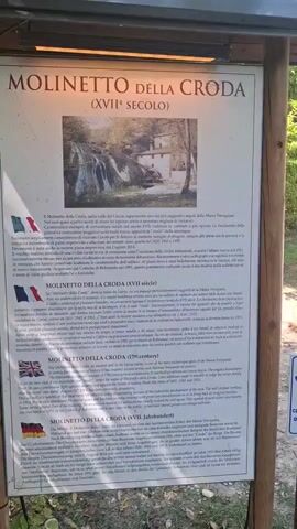 The video begins with a close-up of a sign titled "MOLINETTO DELLA CRODA (XVII SECOLO)". The sign has multiple sections in different languages, indicating historical information about a mill. The first language appears to be Italian, followed by English, and then German. There is a historical photograph on the sign depicting a building and a waterfall. To the right of the sign, a small sign with a dog icon and text "cani al guinzaglio obbligo raccolta deiezione" is visible, which translates to "dogs on a leash, mandatory waste collection".

The camera then pans to the right, revealing a park-like setting with trees, green grass, and a gravel path. In the background, a building complex with a waterfall can be seen. Several people are walking along the path. The first person visible is a woman in a dark top and skirt, carrying a bag and walking towards the camera. Further down the path, a man in a green t-shirt and shorts walks away from the camera, followed by other people. There are also some barrels and potted plants with red flowers in the foreground. The scene is bathed in sunlight, suggesting it is daytime. The environment is natural and appears to be a tourist attraction or a historical site.
