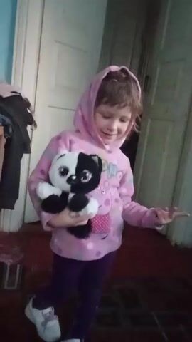 A young girl stands indoors, holding a stuffed panda bear. She wears a pink hooded sweatshirt featuring a Peppa Pig design, purple leggings, and white sneakers. The girl looks at the camera, smiles slightly, and then turns her head downwards briefly. The background features a wooden door, a piece of furniture with clothes draped over it, and a patterned rug on the floor. The lighting appears to be somewhat dim.