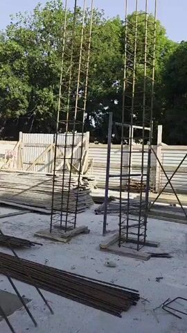 A construction site is shown with two tall rebar cages that are ready to be filled with concrete. In the background, there are trees and a fence. Piles of lumber and rebar are visible in the foreground and on the sides.
