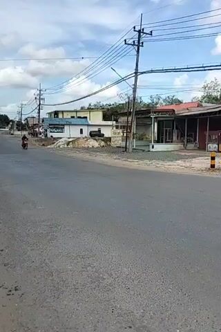 A red scooter with a rider passes by on a paved road. The rider is wearing a dark shirt and a dark helmet. On the right side of the road are several buildings and utility poles with many wires. One building has a red roof and a white front. On the left side of the road, there is a dirt area with some construction materials and a white building. The sky is partly cloudy.