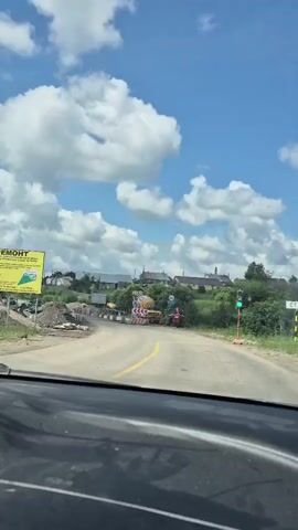 The video shows a view from inside a car driving towards a road construction zone. The sky is blue with white clouds. The road ahead is under repair, with barriers, construction materials, and equipment visible. A cement mixer truck and a red tractor are present. Traffic signs indicating road work and a 30-ton weight limit are also visible. The car appears to be moving slowly towards the construction area.