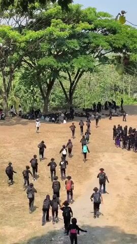 The video shows a group of people, primarily young individuals, in an outdoor area with dirt ground and trees. Many of them are wearing black clothing and carrying backpacks, suggesting they might be students or participating in an organized activity. They are seen walking and moving in various directions across the open space. Some are gathered in groups, while others are walking individually or in smaller clusters. The environment appears to be a natural setting, possibly a field or park, with lush green trees and foliage in the background. The lighting indicates it is daytime, with sunlight casting shadows.