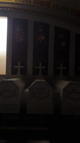 This video shows a dimly lit interior with several sarcophagi in the foreground. Behind the sarcophagi, there are three tall, narrow banners displayed vertically. Each banner has a crest at the top, followed by text and a cross. The sarcophagi are made of white marble and have inscriptions and decorative elements. The floor is marble, and there is a large, brightly lit vertical window or opening to the left of the banners. The overall atmosphere is solemn and appears to be a memorial or a tomb. The text on the banners and sarcophagi suggests a royal or historical context.