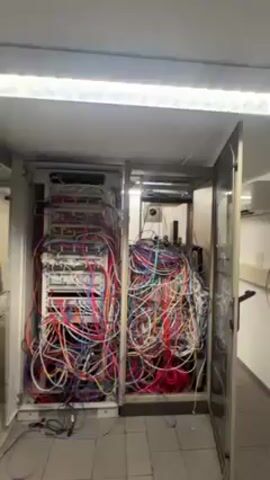 The video begins with a wide shot of two open server cabinets, one on the left and one on the right, both densely packed with wires of various colors. The wires create a tangled, disorganized appearance within each cabinet. The cabinets are lit by a light fixture above them, casting light onto the wires and the surrounding area. The floor appears to be tiled, and a glimpse of a hallway is visible to the right.  The camera then cuts to complete darkness, with only a few blinking green lights visible in the center of the frame, before the video ends.