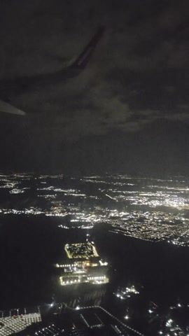 The video shows an aerial view of a brightly lit city at night. Visible in the foreground are the red wingtip and a small portion of the wing of an airplane. The cityscape below consists of numerous lights, appearing as clusters and lines, indicating buildings, streets, and potentially bodies of water reflecting the lights. There are clouds above, partially obscuring the view of the sky.