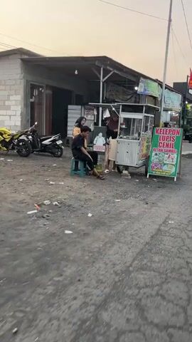 The media file captures a street scene in Kedungcangkring, Indonesia, likely during the late afternoon or early evening, as indicated by the soft, warm lighting. The setting appears to be a modest commercial area with low-rise buildings and a dirt road.

In the foreground, a man is seated on a blue stool on the roadside, looking down. He is dressed in dark clothing. Behind him, a food cart is set up, attended by at least two other individuals. One person, wearing a dark top, appears to be serving food, while another, in a brown shirt, is standing near the cart. A yellow and black motorcycle is parked to the left of the scene, with another white motorcycle parked further back.

The food cart has a prominent sign displaying the text "KELEPON LURIS CENIL KETAN SAMBEL" in green and white lettering. Another sign above the cart is partially visible, showing text that might relate to "LUEKOR". The area around the cart and road is dusty and somewhat unkempt, with scattered debris.

The overall atmosphere seems relatively calm, with a focus on the street food vendor activity. The lighting suggests the sun is low in the sky, casting long shadows and contributing to a golden hour feel. The weather appears to be clear. The presence of the food cart and the various vehicles suggests a place of local commerce and daily life.