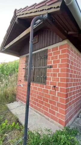 The video pans across the exterior of a small, brick building with a wooden roof and siding. The building has a window with wooden shutters and a metal pipe running vertically along the side. There is also a door visible on the right side of the building. The area around the building is overgrown with grass and other vegetation.