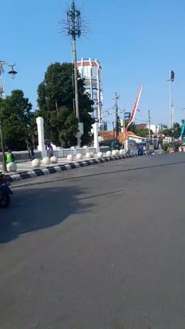 The video shows a street scene with a tall, cylindrical building in the background, possibly a water tower or a communication tower, painted with white and red vertical stripes and blue accents. A flag, predominantly red and white, is mounted on a pole near the tower. In the foreground, there is a paved road with a black and white striped curb. Several small stalls or carts are set up along the sidewalk, with some people attending them. A motorcycle with two riders is moving on the road. The environment appears to be an urban or semi-urban setting with a clear blue sky.
