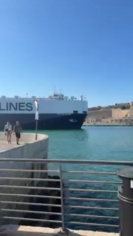 The video shows a large cargo ship with "NEPTUNE LINES" written on its side moving on the water. A couple is walking on a concrete pier beside the water. The camera is positioned behind a railing and a concrete structure, capturing the scene with a clear blue sky in the background.