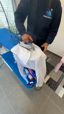A person wearing a dark blue sweatshirt with a logo that says "StrijkExpert" is ironing a white shirt on a blue ironing board. The person is using a silver and black steam iron. Steam is visible coming from the iron and the shirt. There are several bottles of cleaning products on the ironing board beneath the shirt. The ironing board is set up in a room with light-colored tiled floors and a window with blinds.