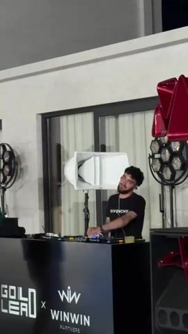 A male DJ with dark hair and a beard is seen from the waist up, wearing a black t-shirt. He is standing behind a black DJ booth with logos that read "GOLD LEAD x WINWIN PARTNERS". He is operating a DJ mixer. Two large speakers, one white and one red, are visible on either side of him. The background shows a building with large windows and curtains. The lighting is dim, suggesting an evening or indoor event. The DJ appears to be focused on his equipment.