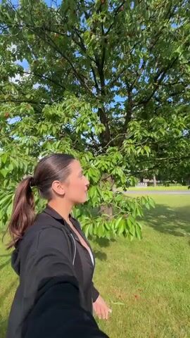 A young woman with dark hair tied in a low ponytail, wearing a black zip-up jacket over a black top and a white undershirt, holds a camera in selfie mode. She stands on green grass with a large leafy tree bearing red fruits directly behind her. In the background are more trees, a paved area, and a clear blue sky. The woman initially faces the camera, smiling. She then slowly turns her head to her right, looking off into the distance, before turning back to face the camera and smiling again.