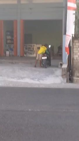 At 00:00, a person wearing a yellow shirt is seen on a motorcycle in front of a building with orange doors. They appear to be interacting with the motorcycle. At 00:01, the person is still with the motorcycle, and the motorcycle's taillight is illuminated. By 00:02, the person has moved further into the building's entryway, and the motorcycle is still present. At 00:03, the person is further inside the building, and the motorcycle is now positioned more towards the rear. By 00:04, a different motorcycle with two people on it rides past in the foreground, out of focus, while the person in the yellow shirt is no longer visible within the building's entrance. A flag with red and white stripes is visible on the right side of the frame. The environment appears to be an outdoor area in front of a commercial building.