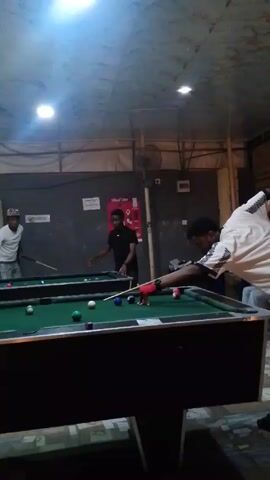 Three men are playing pool in what looks like a casual billiards hall.  The room has two pool tables with green felt, and the tables have black frames.  The lighting is provided by two or three bright ceiling lights.  The walls are a drab grey color and adorned with advertisements for alcohol. One man, in the foreground, is wearing a light-colored patterned shirt. He is in the middle of taking a shot. Another man, standing near the other table, is wearing a black t-shirt. The third man, standing behind him, is wearing a white t-shirt. All three men appear to be relatively young. The room has a casual, relaxed atmosphere.  There is music playing, discernible by a heavy beat, which indicates that this may be a public venue.
