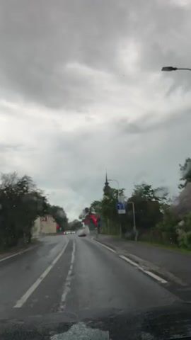 The video shows a view from inside a vehicle, looking through a rain-streaked windshield at a road ahead. It is a cloudy, overcast day, and the road is wet, indicating rain. Trees line both sides of the road, and buildings are visible on the left. Traffic lights are visible, one of which appears to be red. Several cars are on the road, moving in the same direction as the camera. The overall impression is one of driving in wet weather.