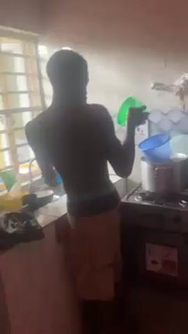 A shirtless man with dark skin is washing dishes in a kitchen. He is wearing khaki shorts and underwear. There is a window with bars behind the sink. The sink area is cluttered with dishes, containers, and a trash bag. A stove with pots and pans on it is visible in the background. The man is focused on washing a blue bowl in the sink.