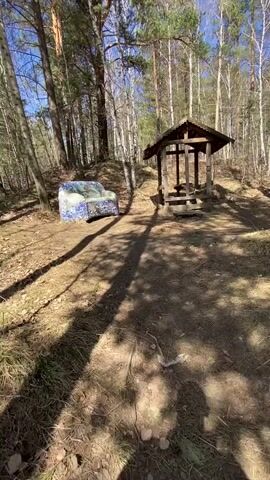 The camera is aimed at a dirt path in a forest. To the left of the path is a mosaic-covered armchair. To the right of the path, further in the distance, is a small wooden gazebo. The scene is lit by bright sunlight that casts long shadows. The trees are mostly bare, with some green pine trees visible. The ground is covered in leaves and small bits of debris. The sky is blue and clear.