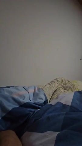 From a low angle in a dim room, a person is shown lying in bed, partially covered by a blue and white patterned blanket. A hand with blue painted nails emerges from under the blanket and touches another arm, which is giving a thumbs up. The background is a plain, light-colored wall.