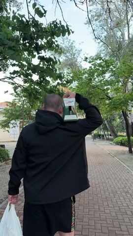 A man walks away from the camera on a brick pathway flanked by trees. He carries a white plastic bag in his left hand. On his right shoulder, he balances a six-pack of San Miguel Especial beer. The man is bald and wears a black hooded jacket. The scene appears to be in a park setting with scattered trees and a lamppost visible.
