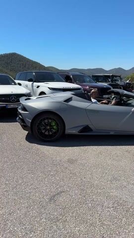 The video shows a gray Lamborghini Huracan convertible parked on an asphalt road, featuring a driver wearing a light-colored shirt. Green brake calipers and black rims are visible on the car. A Mercedes-Benz, Range Rovers, and other vehicles are parked behind and beside the Lamborghini. The backdrop includes a clear blue sky, mountains, and trees. People are seen in the background, some standing near the vehicles. The camera pans slightly to the right, revealing a black Porsche parked behind the Lamborghini. The Lamborghini driver is shown sitting in the driver's seat. There is an audible engine rev from the Lamborghini.