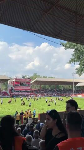 The video captures an outdoor wrestling event with a crowd gathered in a stadium. The field is covered in green grass, and multiple wrestlers, most wearing white shirts and dark pants, are engaged in matches. Spectators fill the bleachers under a red canopy and around the field. A large banner with Turkish writing is visible on a building behind the field. The sky is blue with scattered white clouds, and trees are present in the background. The video appears to be filmed from the perspective of someone in the audience.