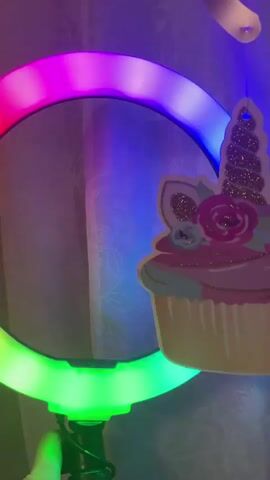 A hand is holding a ring light that is lit with multiple colors. A pink unicorn cupcake decoration is hanging from the top right of the ring light. The background appears to be a wall with a patterned curtain. The ring light is turned on and cycling through a rainbow of colors.