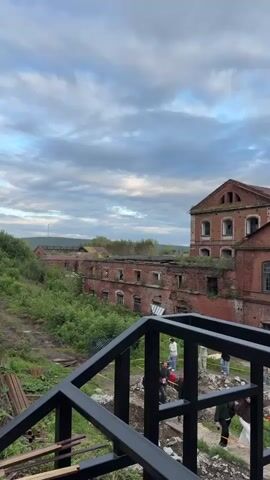 The video pans across a partially constructed outdoor area in front of a derelict red brick industrial building. In the foreground, a black metal railing and frame suggest ongoing construction. Beyond this, the abandoned building, with its broken windows and overgrown vegetation, dominates the scene. Several people are visible in the middle ground, standing near the building and what appears to be a trench or excavation. The sky is cloudy, with patches of blue.